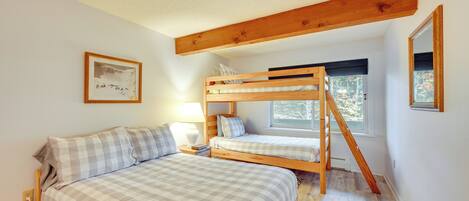 A bedroom with a bunk bed, a single bed, a bedside table with a lamp, a framed picture on the wall, and a window with a view of trees.