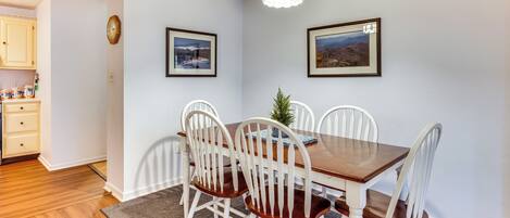 A dining area with a wooden table and white chairs, a small plant, and two framed pictures on the wall.