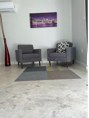 Um quarto com duas poltronas cinzentas, um vaso vermelho, um quadro de paisagem urbana e um tapete cinzento.