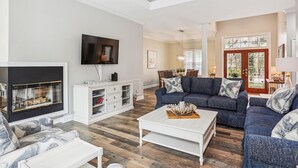 A spacious living room with a large flat-screen TV, a white entertainment unit, a blue sofa set, and a dining area in the background.