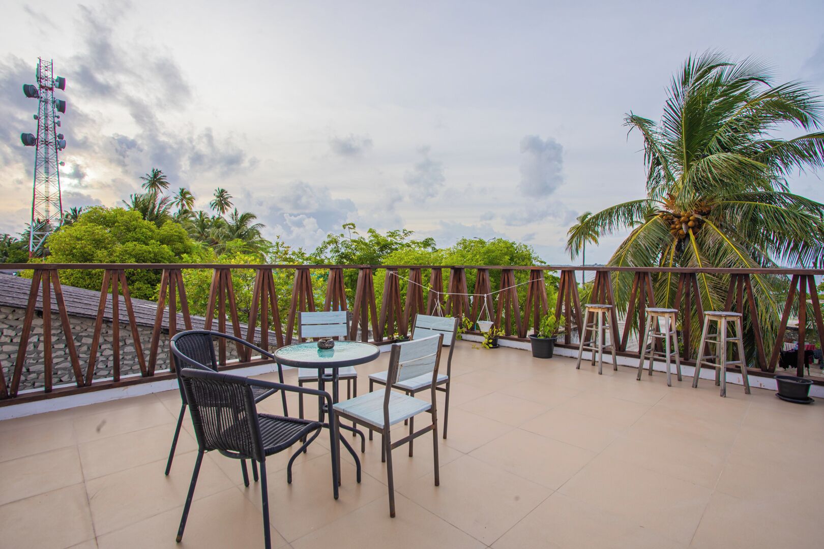 A rooftop terrace with a table and chairs, surrounded by a wooden railing, palm trees, and a clear sky.