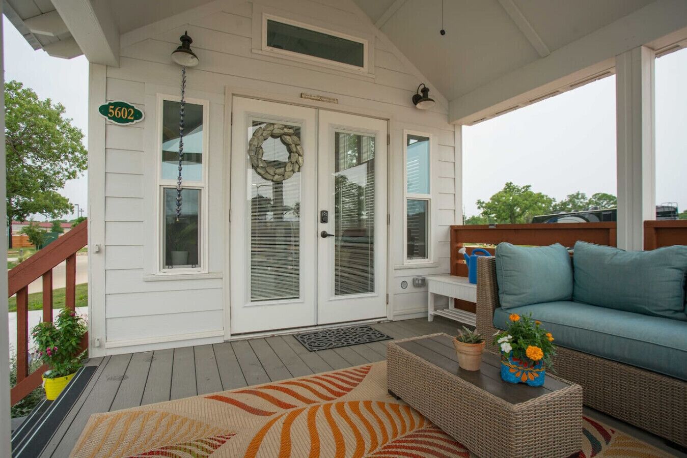 A covered porch with a white exterior, a number sign '5602', a stone wreath on the door, a blue couch, and a wicker table with a vase of flowers.