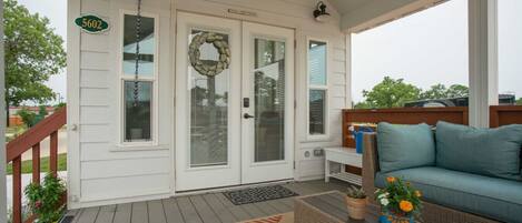 A covered porch with a white exterior, a number sign '5602', a stone wreath on the door, a blue couch, and a wicker table with a vase of flowers.