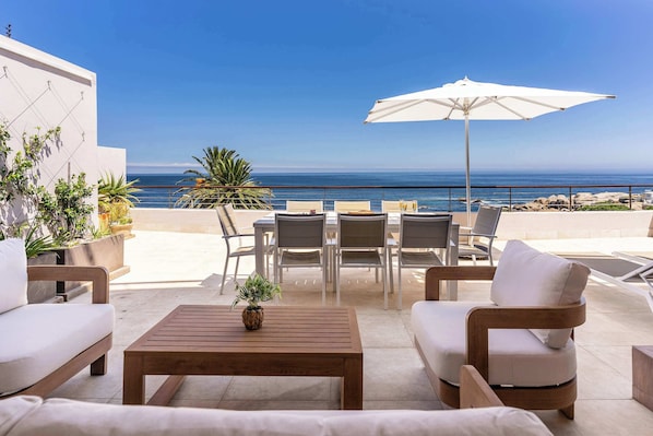 Un espace extérieur aménagé avec des sièges, offrant une vue sur l’océan, comprenant une table en bois, des canapés blancs et un grand parasol.