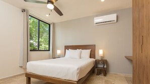 Un dormitorio con cama, dos mesitas de noche con lámparas, ventilador de techo, equipo de aire acondicionado y ventana con cortinas.