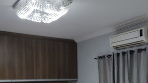 Une chambre à coucher comprenant un lit, une tête de lit capitonnée, un luminaire au plafond, un appareil de climatisation et un rideau.