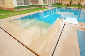 Una piscina con fondo de vidrio transparente, rodeada por una solana con baldosas y un césped bien cuidado.