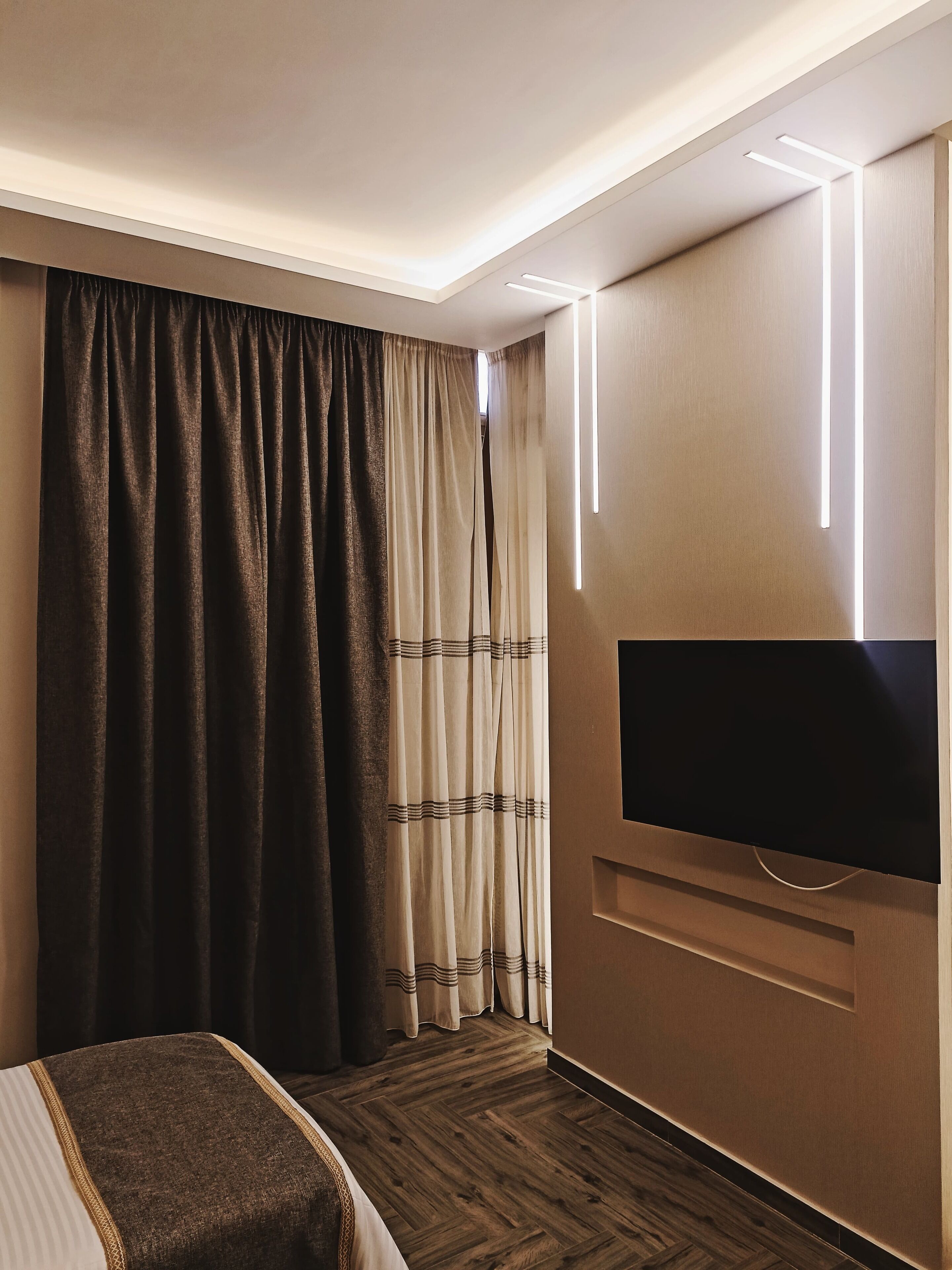 Habitación de hotel con cama, televisor de pantalla plana montado en la pared y un ventanal con cortinas.