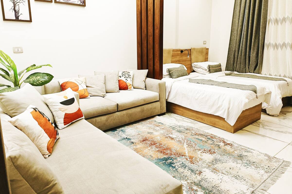 Una habitación de hotel moderna con un sofá, una cama, un cabecero de madera y un amplio ventanal con cortinas.