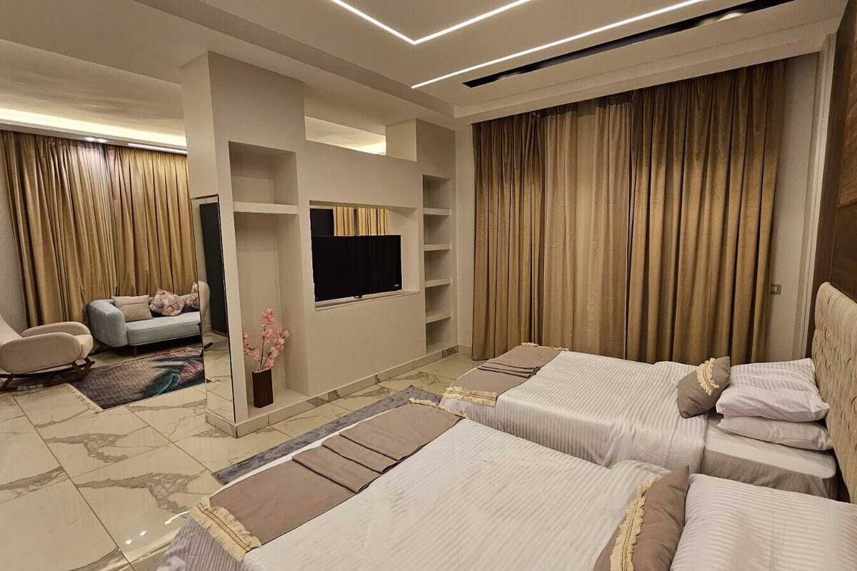 Una habitación de hotel moderna con dos camas, un sofá, una silla, un televisor y un amplio ventanal con cortinas.