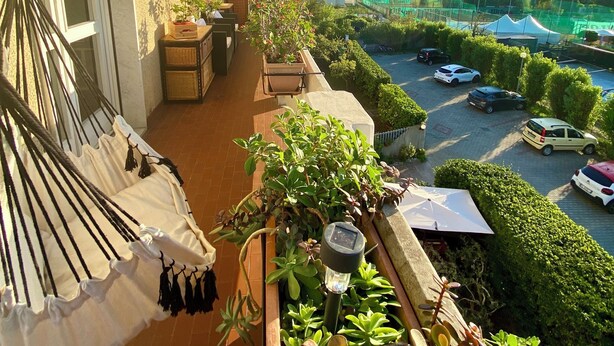 Un balcón con una hamaca, plantas en macetas y vistas a un aparcamiento y montañas.