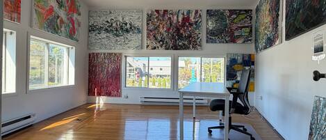 A spacious room with large windows, a white desk, and multiple abstract paintings on the walls.