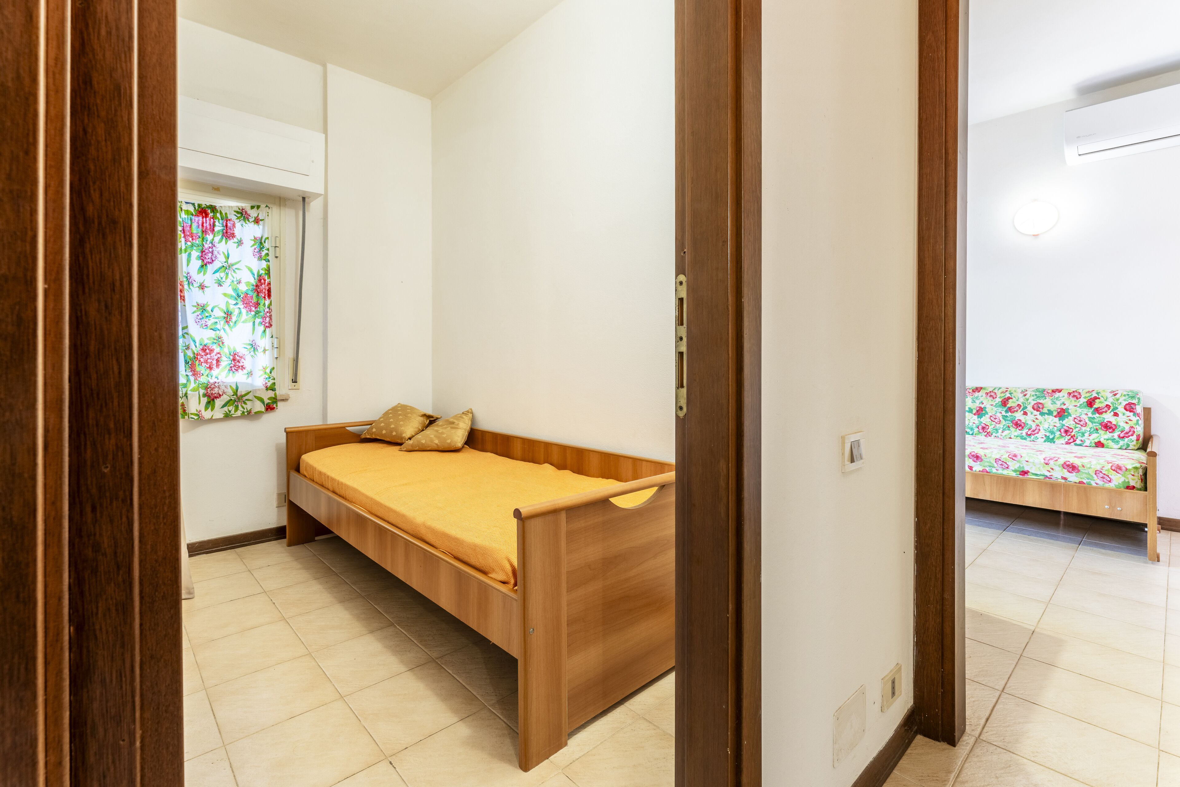 Een kleine eenpersoonskamer met een houten bed, een raam met bloemen gordijnen, en een betegelde vloer.