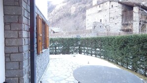 Un edificio de piedra con contraventanas de madera, un patio de piedra y un seto.