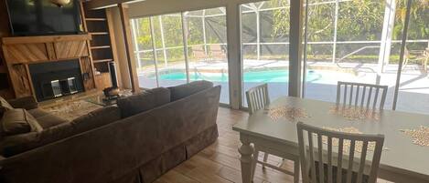 A spacious living area with a wooden ceiling, a ceiling fan, a fireplace, a large window overlooking a pool, and a dining table with chairs.