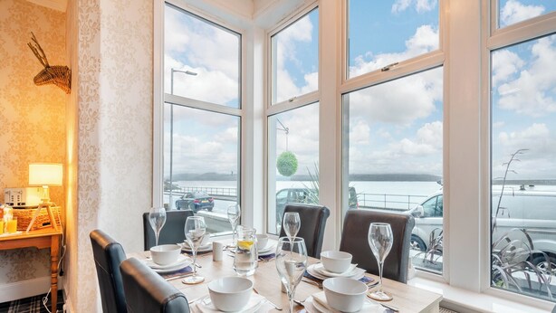 Un coin repas avec une table dressée pour quatre personnes, de grandes fenêtres offrant une vue sur la mer et les voitures garées, et un élément décoratif mural.