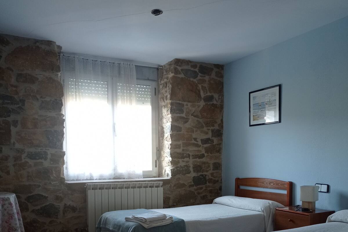 A room with two beds, a stone wall, a window with curtains, a radiator, and a framed picture on the wall.