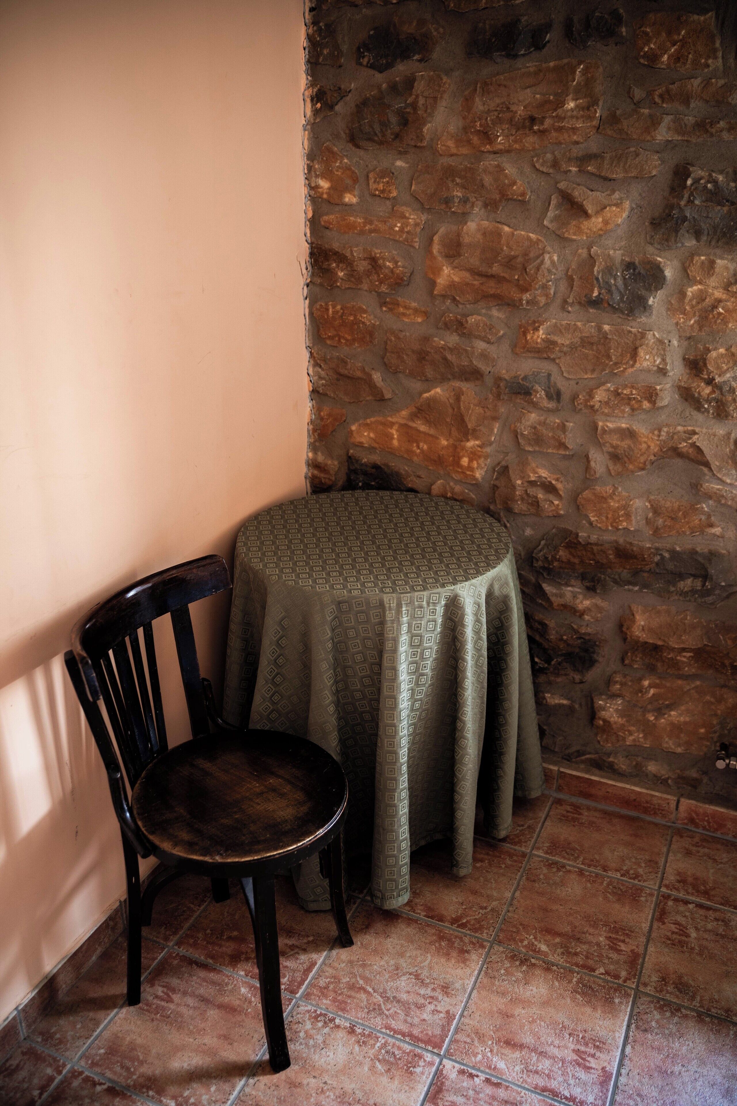 A corner with a stone wall, a round table with a patterned tablecloth, and a wooden chair.
