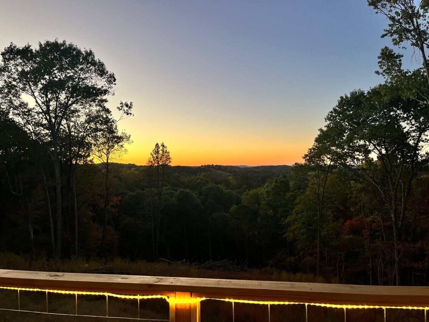 A sunset view from a balcony with string lights, surrounded by trees.