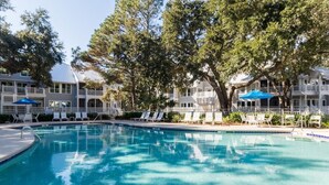 A swimming pool with lounge chairs and umbrellas, surrounded by trees and a building with balconies.