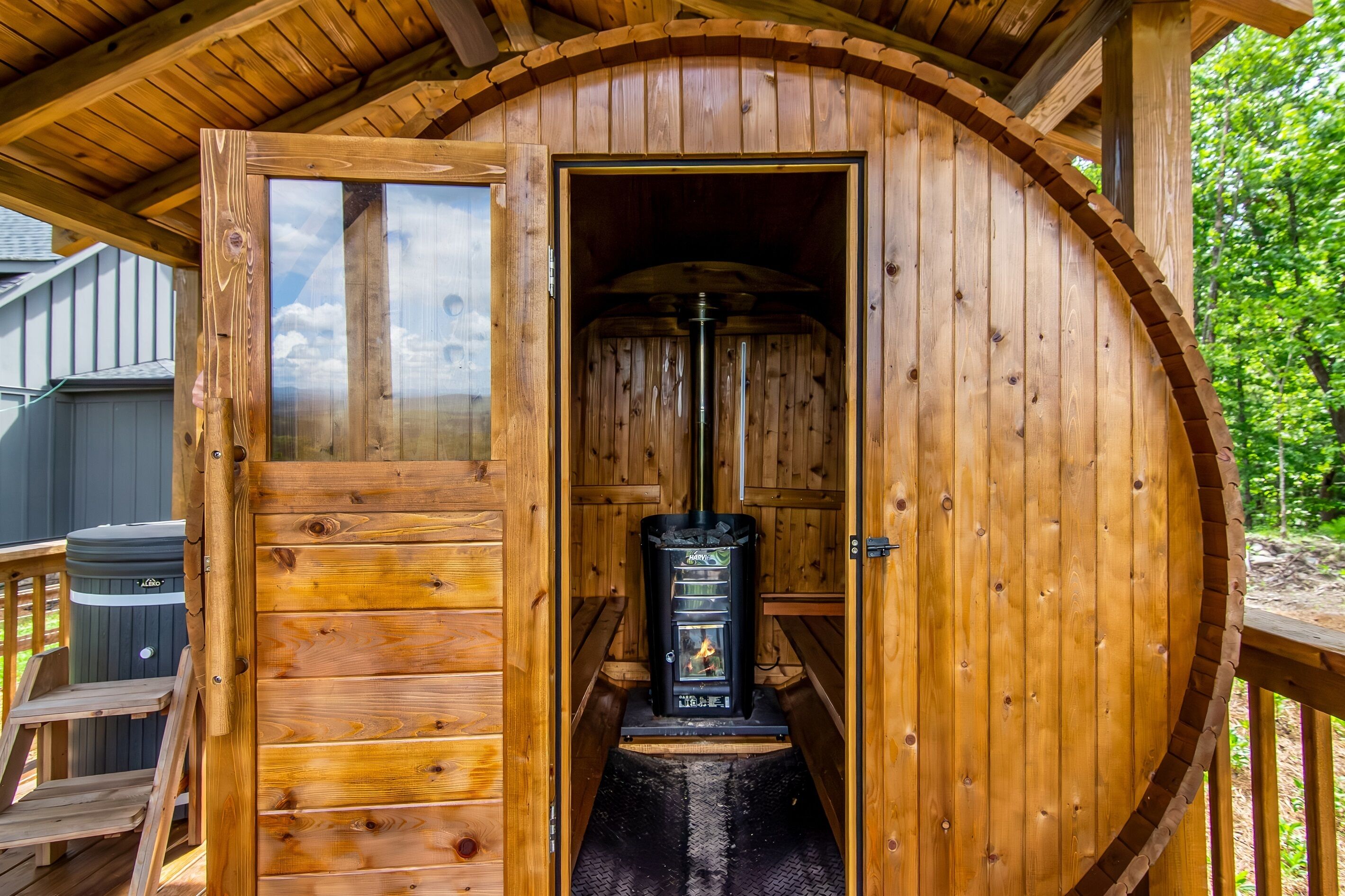 A wooden sauna with a glass door, a black stove, and a wooden deck outside.