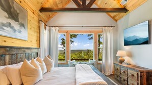 A bedroom with a large bed, a wooden headboard, a wooden dresser, and a view of greenery through large windows.