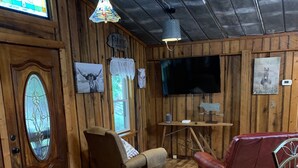Ein rustikaler Innenraum mit Holzwänden, Holzboden, einem an der Wand montierten Fernseher, einem roten Ledersofa, einem braunen Sessel und einer Holztür mit Buntglasfenster.