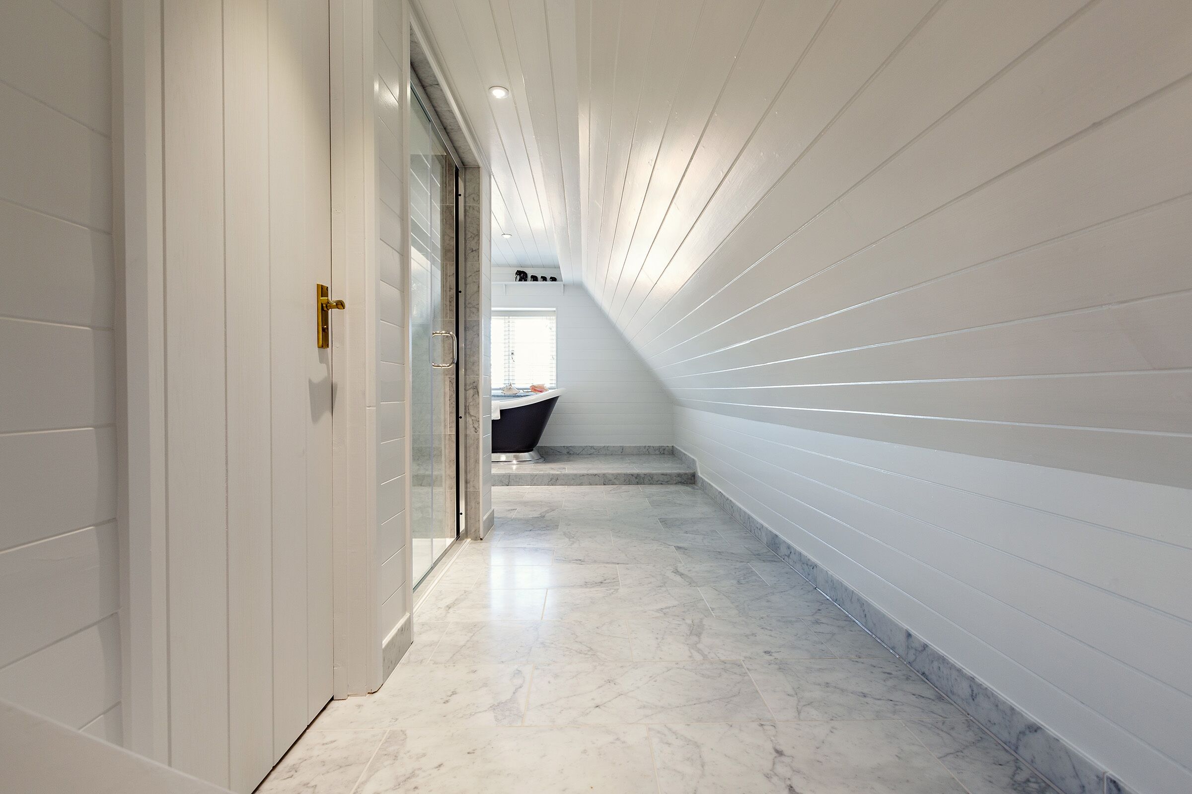 Un pasillo moderno con paredes blancas y suelo de mármol, que conduce a un baño con bañera negra y ventana.