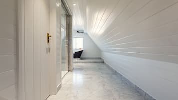 Un pasillo moderno con paredes blancas y suelo de mármol, que conduce a un baño con bañera negra y ventana.