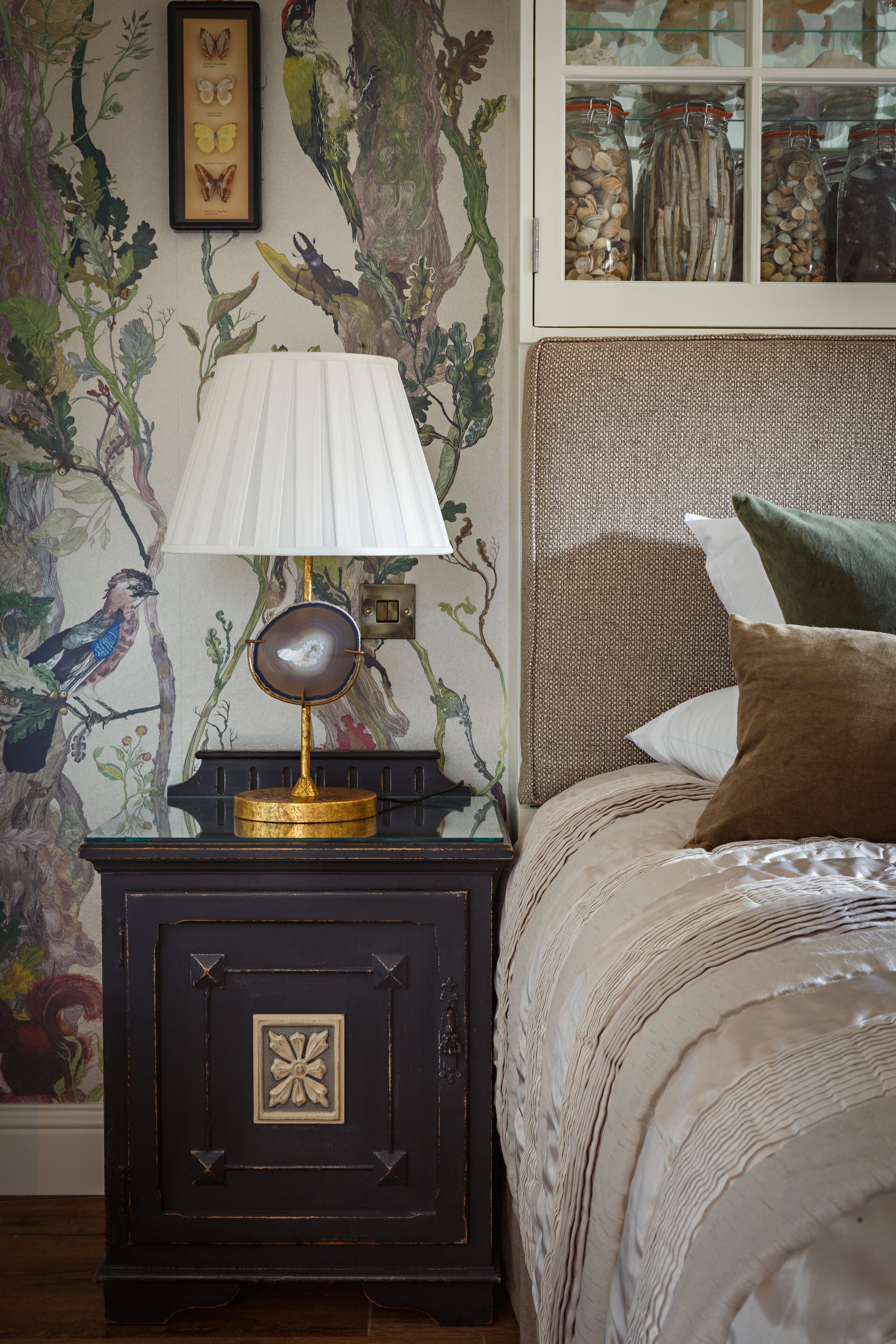 A bedroom with a bed, a nightstand with a lamp, and a wall with a nature-themed mural.