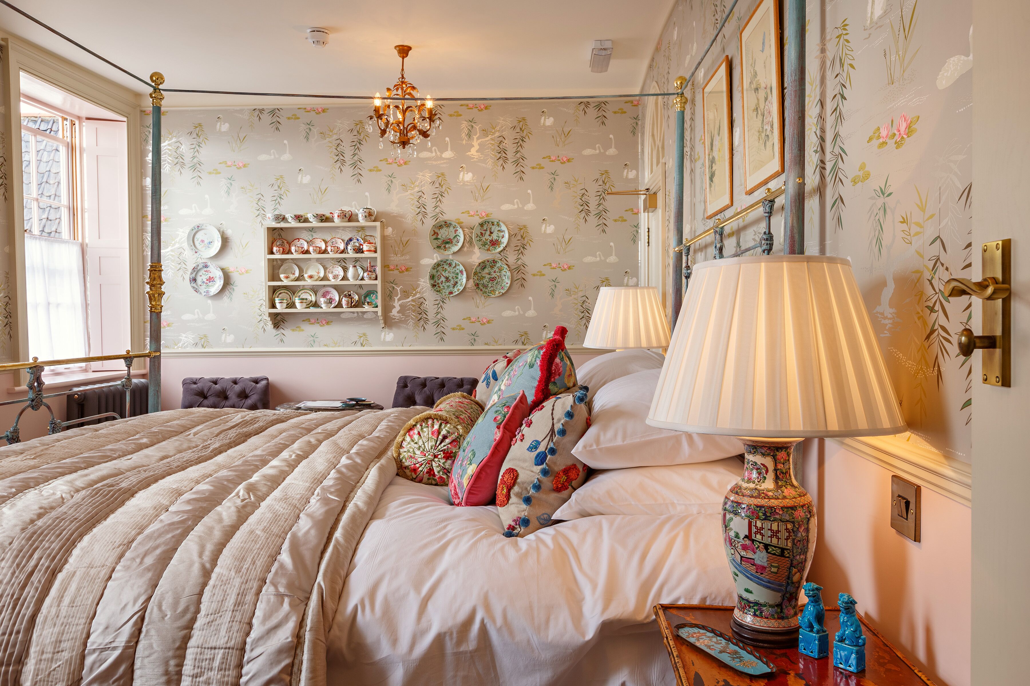Une chambre à coucher avec un grand lit, des coussins décoratifs, une lampe de chevet et des étagères murales où sont posées des assiettes.