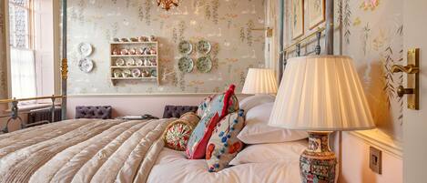 Une chambre à coucher avec un grand lit, des coussins décoratifs, une lampe de chevet et des étagères murales où sont posées des assiettes.