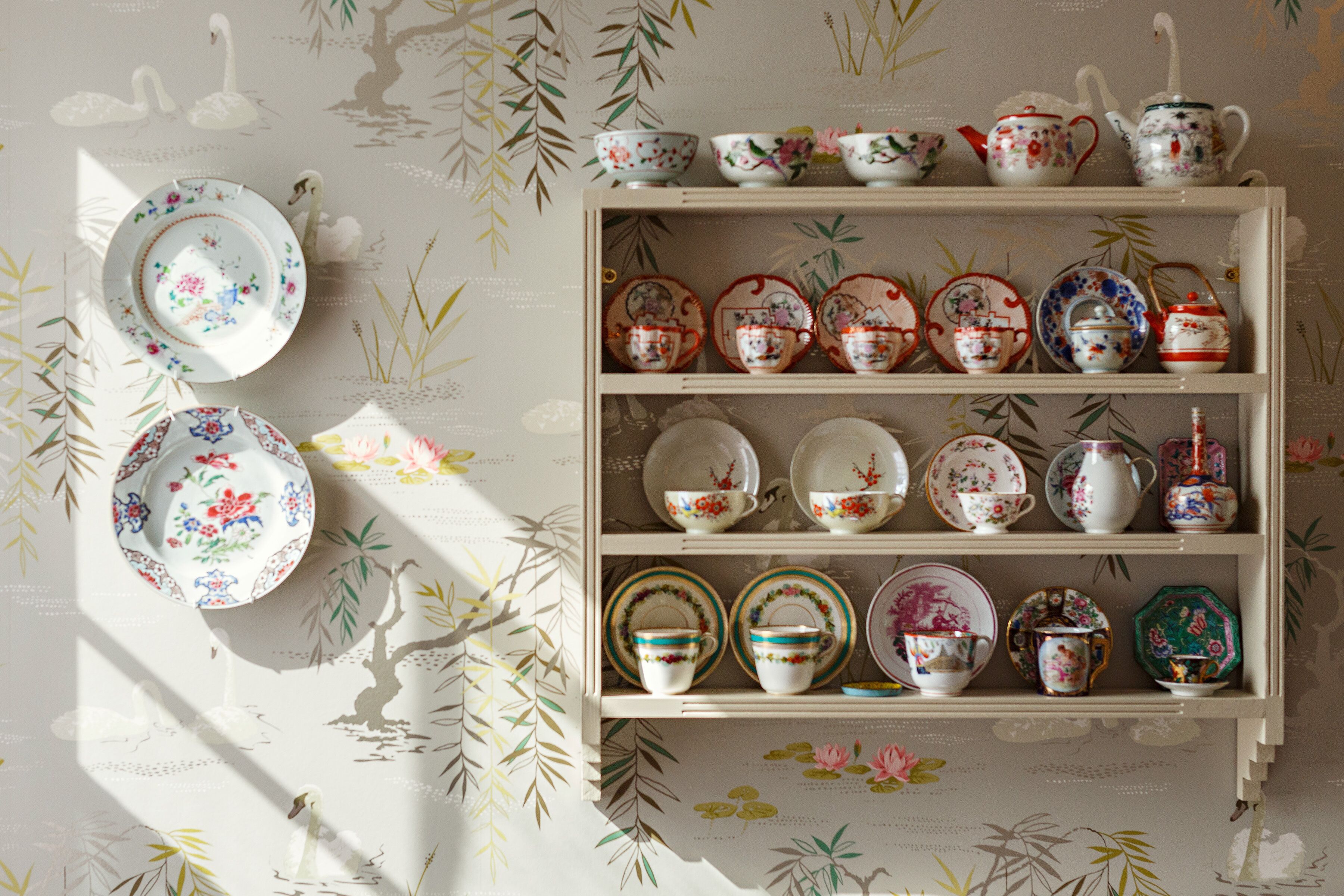 A display of various ceramic tea sets and plates arranged on shelves against a wall with a floral wallpaper pattern.