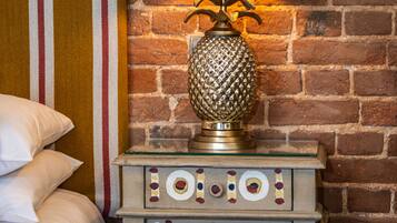 A bed with pillows, a striped blanket, a lamp with a yellow shade, and a decorative cabinet against a brick wall.