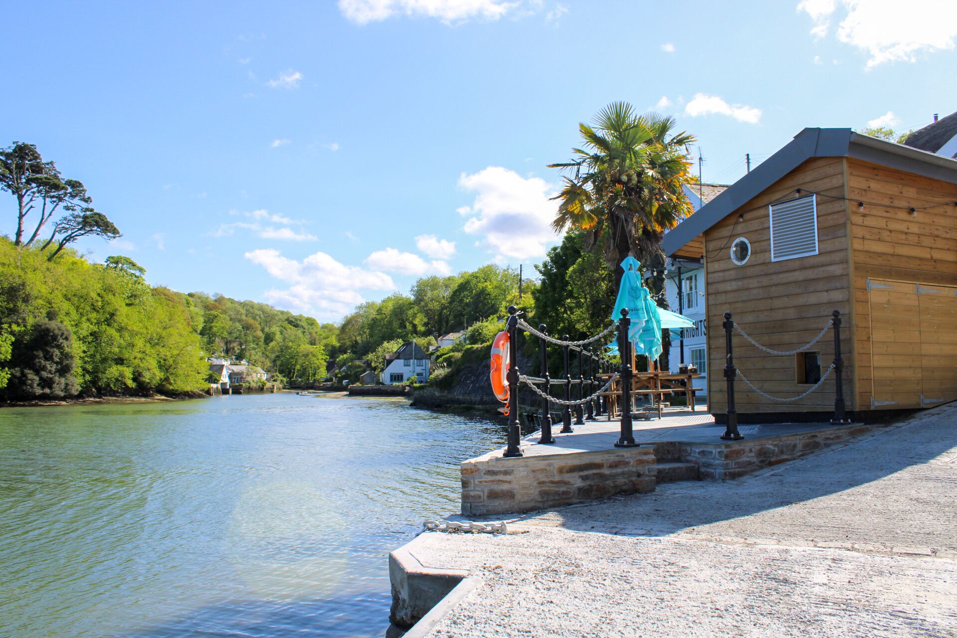 A wooden building with a stone dock extending into calm water, surrounded by lush greenery and a clear sky.