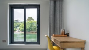 A room with a large window overlooking a body of water and greenery, a wooden desk with a chair, and a grey curtain. - The Shipwrights Arms (Helston)