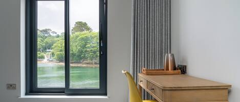 A room with a large window overlooking a body of water and greenery, a wooden desk with a chair, and a grey curtain.