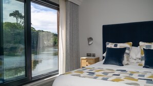 A bedroom with a large window, a bed with patterned bedding, a nightstand, and a wall-mounted lamp. - The Shipwrights Arms (Helston)