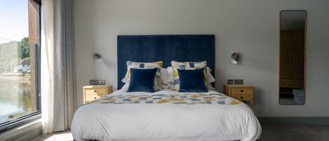 A modern bedroom with a large bed, a nightstand, a mirror, and a view of the outdoors.