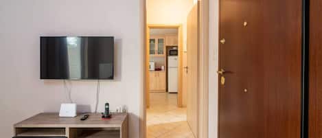 A room with a TV mounted on the wall, a wooden desk with a drawer, a door, and a kitchen visible through an open door.