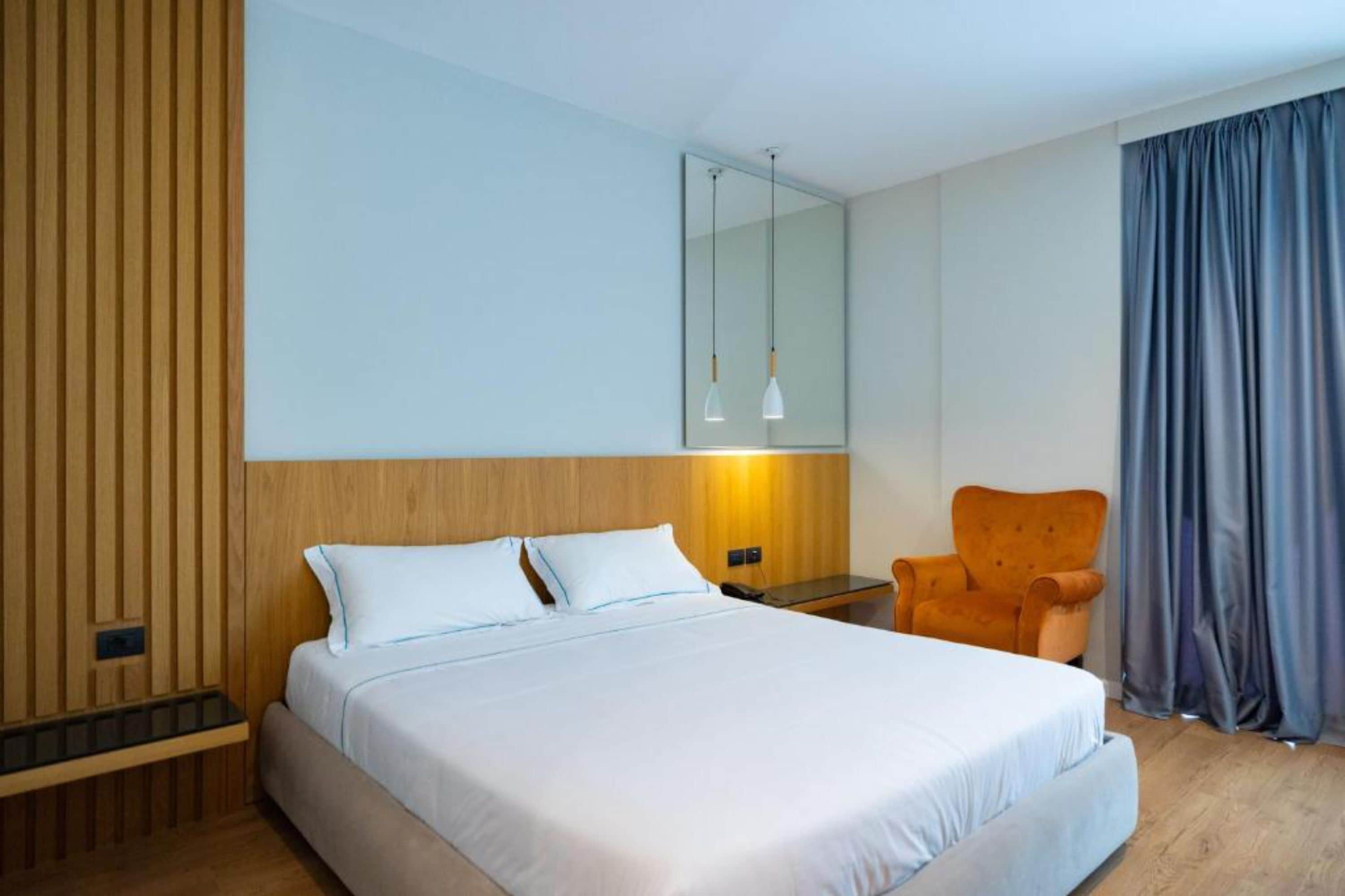 A modern hotel room with a large bed, a wooden headboard, a small table, an orange armchair, and a large window with curtains.