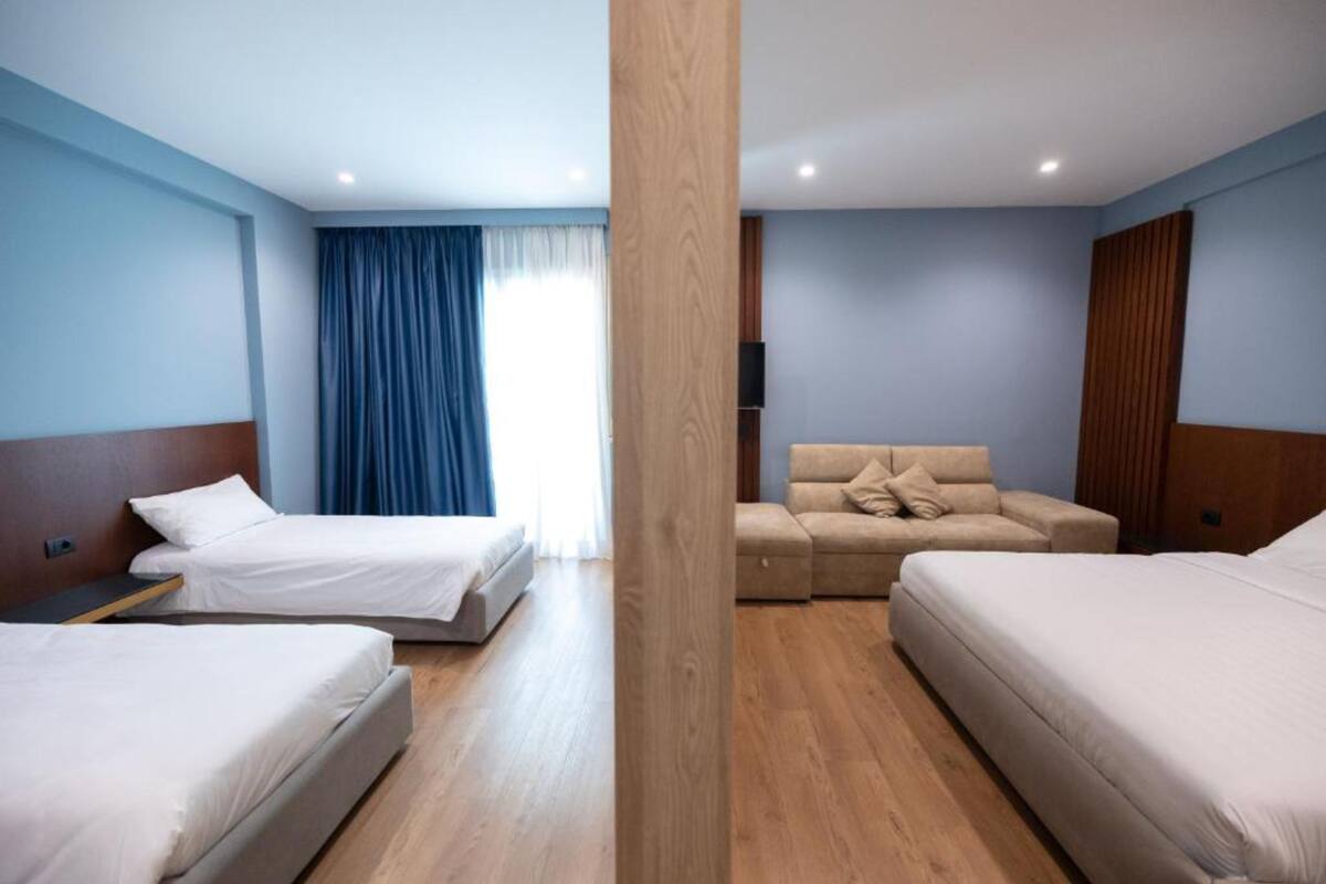 A hotel room with two single beds, a sofa bed, wooden flooring, and a large window with curtains.