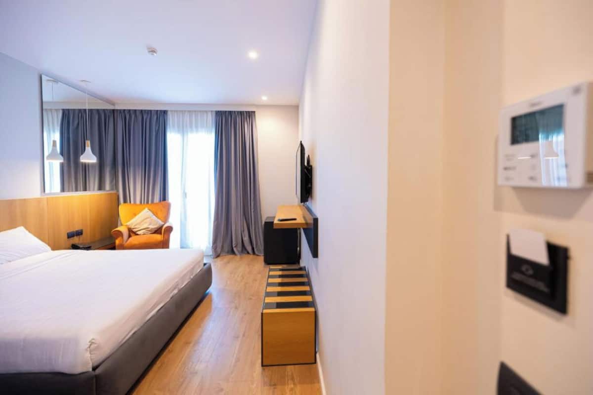 A hotel room with a bed, a chair, a desk, a standing lamp, a thermostat, and a window with curtains.