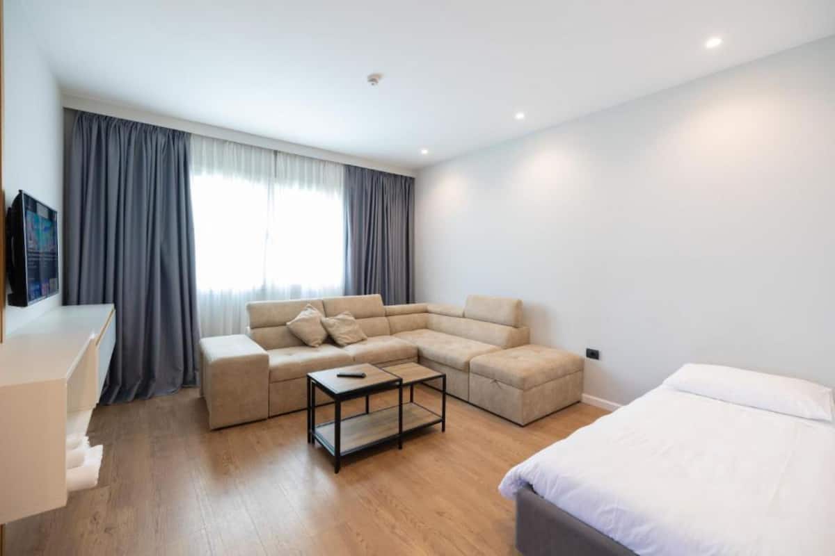 A modern, well-lit room with a sectional sofa, a small coffee table, a bed, and a TV on a stand.
