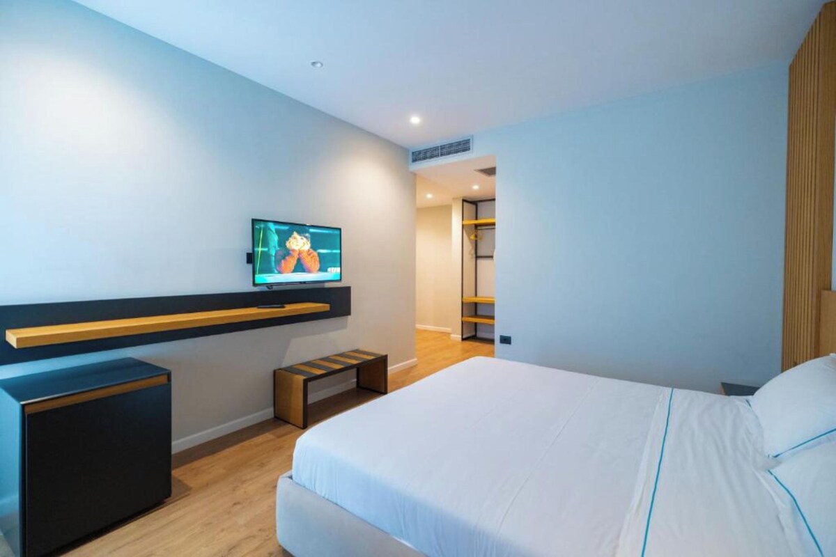 Una habitación de hotel moderna con una cama, un televisor instalado en la pared, un banco de madera y un estante empotrado.
