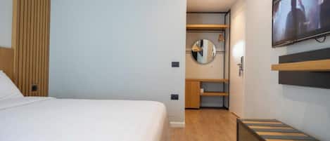 A modern hotel room with a large bed, a TV mounted on the wall, a wooden wardrobe, and a small seating area.