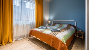 Ein ordentlich gemachtes Bett mit einer farbenfrohen Steudecke, ein Nachttisch mit einer Lampe und ein Fenster mit gelben Vorhängen.