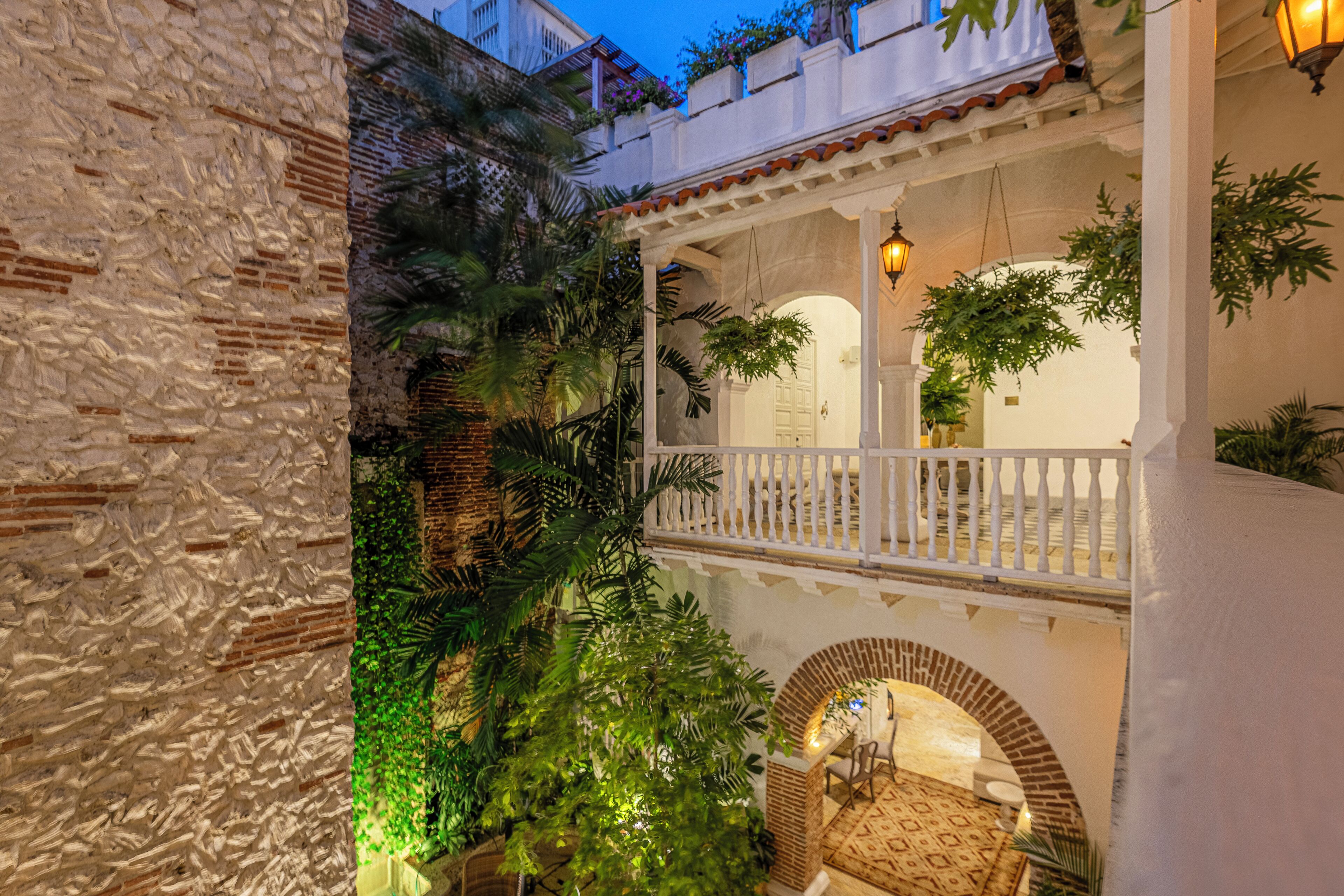 Un patio con balcón blanco, pared de ladrillo y abundante vegetación.