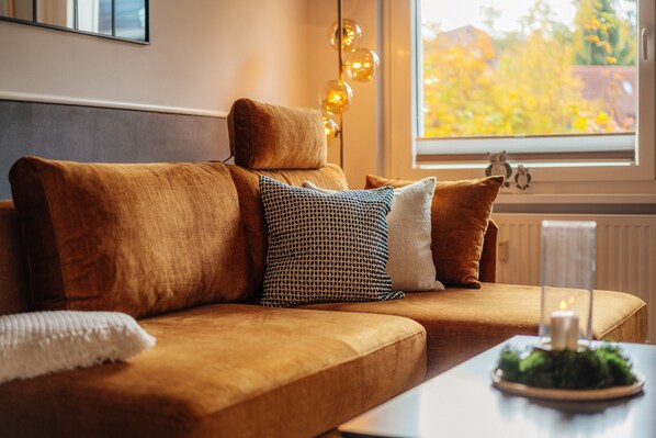 Een gezellige woonkamer met een bruine bank, kussens met een patroon, een witte plaid en een brandende kaars op een tafel.