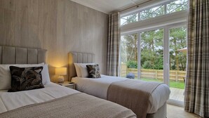 A bedroom with two beds, a nightstand with a lamp, and a view of the outdoors through large windows. - Elderberry Lodge | sleeps 4 - Hot Tub, Dog Friendly - 5* Gold Award (Pickering)